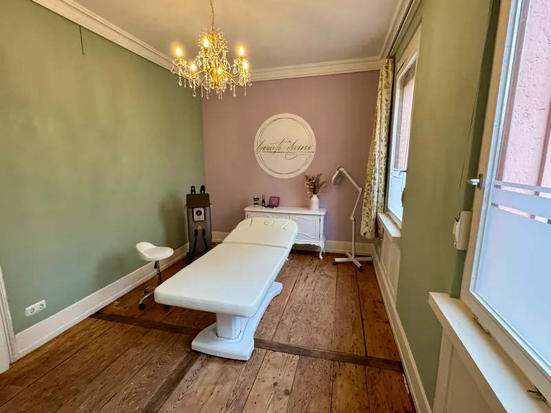 Behandlungsraum im Studio Stuttgart mit Liege, Kronleuchter und dem Logo von beauté divine an der Wand